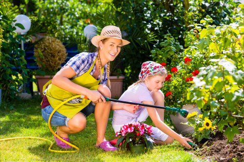 A woman and a young girl are gardening together in a lush, well-maintained backyard garden, with vibrant flowering plants and dense greenery in the background. The woman, wearing a straw hat, a checkered shirt, and a yellow apron, is crouching and holding a garden hose, smiling as she assists the girl. The girl, dressed in a light-colored outfit and a floral hat, is kneeling on the grass, carefully planting or tending to flowers in a flower bed bordered by a variety of colorful blossoms, including reds and yellows. The lawn is neatly trimmed with lush green grass, and the garden features a mix of flowering shrubs and leafy plants, creating a lively and inviting outdoor environment suitable for family outdoor activities and garden care. Soft sunlight filters through the trees, illuminating the scene with a warm, natural glow, emphasizing the peaceful, productive atmosphere for gardening services in Yiewsley and the broader West London area serviced by Gardening Services Yiewsley.