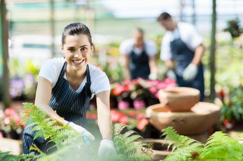 A smiling woman dressed in a striped apron and white t-shirt is engaged in gardening inside a greenhouse, surrounded by vibrant green fern plants and soil. Behind her, two other gardeners, a man and a woman, are working among colourful flowering plants, with the man tending to potted blooms and the woman assisting amidst the lush surroundings. The greenhouse environment features a bright, natural light filling the space, highlighting the healthy, well-maintained plants and the textured soil beds. In the background, there are some stone or terracotta water features, adding decorative and functional elements to the garden scene. The setting demonstrates a neatly organized, productive space that could benefit from professional outdoor maintenance and gardening services in Yiewsley, suitable for both landscaping and plant care, emphasizing a professional and natural outdoor environment typical of a well-kept garden or nursery in the local area.