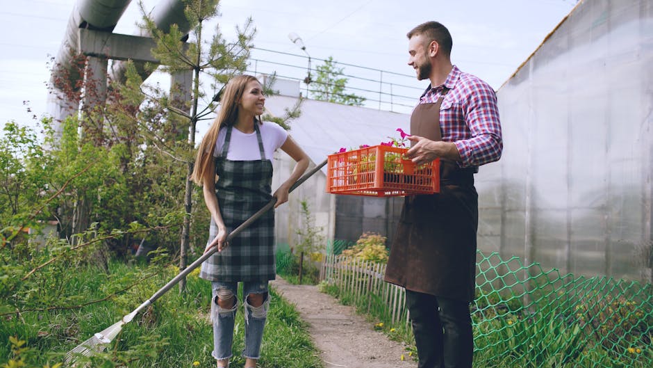 A young woman and a man are engaged in garden maintenance in a lush outdoor setting. The woman, dressed in a white t-shirt, ripped jeans, and a checkered apron, is holding a garden rake and standing o